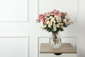 Vase with beautiful flowers on wooden nightstand near white wall, space for text