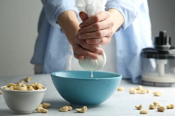 Woman straining cashew milk into bowl at light table with nuts indoors, closeup