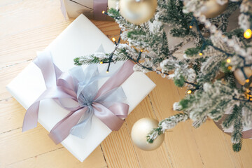 Beautifully wrapped white gift box with bow under Christmas tree decorated with sparkling ornaments and frosted branches, creating a cozy holiday atmosphere.