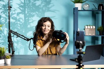 Smiling tech vlogger reviewing VR goggles in living room studio. Happy woman filming virtual reality headset review vlog for gadget enthusiasts using mobile phone and mic