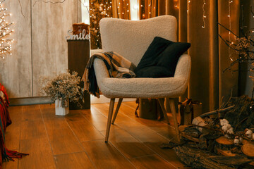 A cozy interior with a soft armchair, a blanket and pillows against the background of a warm garland of lighting. Elements of winter decor create an atmosphere of coziness and celebration