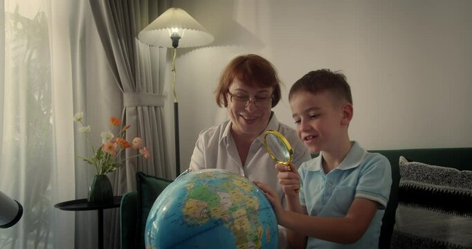 Child with grandmother is studying while sitting at home in rays sunset in room.Kid in morning light sunset, Cute boy Child with Globe and magnifying glass, technology for schoolboy, child looks globe