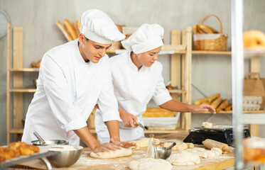 Two young bakers prepare dough in kitchen of bakery - together they knead dough, cut it with a knife and weigh it on scales