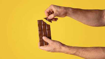 Closeup view of man breaking piece of chocolate from bar, orange background. Copy space. Panorama