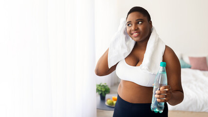 Plus size young African American woman with bottle of water wiping sweat with towel after home...