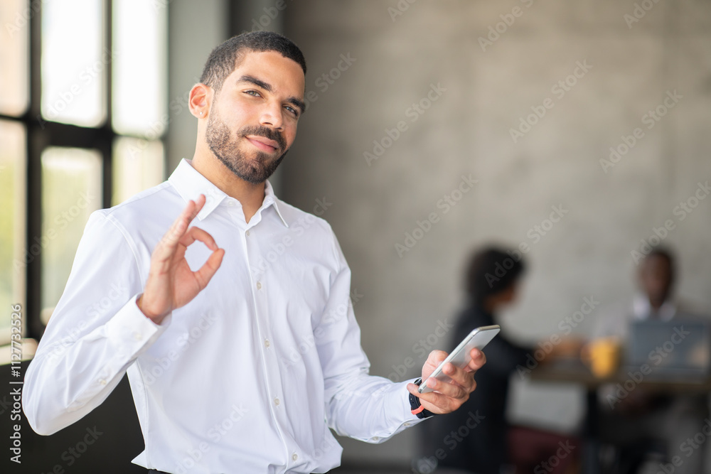 Poster A professional man in a white shirt holds a smartphone while making an 'OK' gesture. He appears confident and engaged in a collaborative office environment with colleagues working in the background.