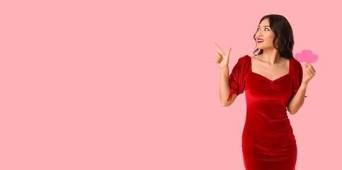 Beautiful happy Asian woman with paper hearts pointing at something on pink background with space for text. Valentine's Day celebration