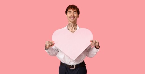 Handsome tattooed young man holding paper heart on pink background. Valentine's Day celebration
