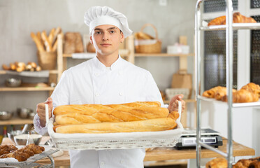 Positive young guy wearing pinafore holding baguettes in square wicker basket in bakery shop