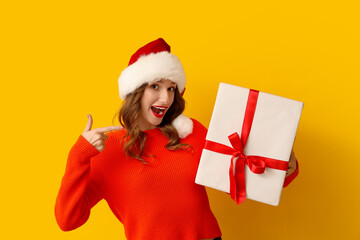Young woman in Santa hat holding Christmas gift box and showing thumb-up gesture on yellow background