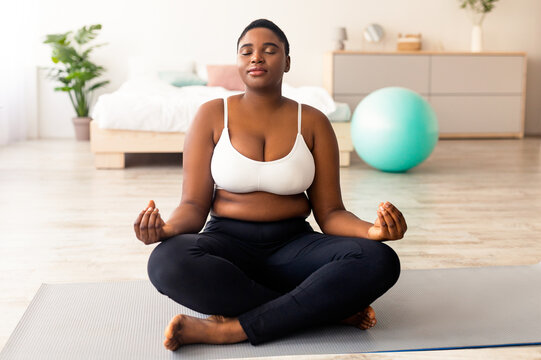 Plus size black woman sitting in lotus pose, meditating with closed eyes, practicing yoga at home. Overweight young lady doing breathing exercises. Wellness, mindfulness concept