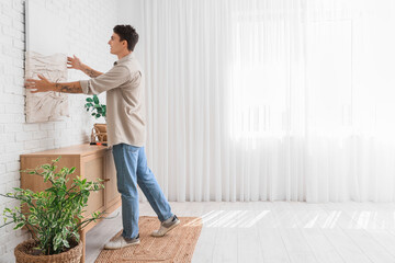 Young man hanging painting on white brick wall at home