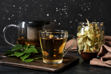 Wooden board with cup of tasty linden tea and leaves on black background