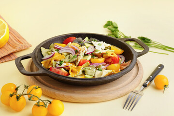Frying pan with delicious Italian pasta salad on light yellow background