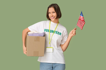 Young female volunteer holding USA flag and cardboard box with clothes on green background