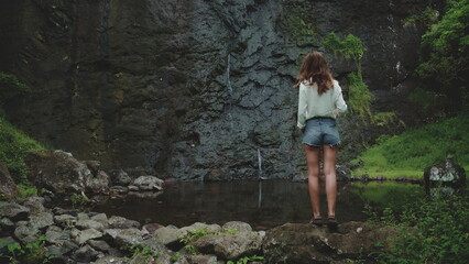 Woman tourist gazes at a waterfall from a cliff in a lush forest, embracing nature's beauty and tranquility