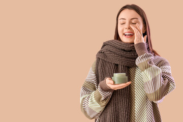 Young woman in winter clothes applying facial cream on beige background