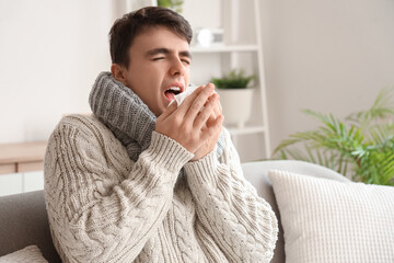 Ill young man with tissue sneezing on sofa at home