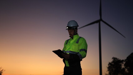 Site engineer with green reflective safety jacket and headlamp working on a digital tablet at a windmill farm © naraichal