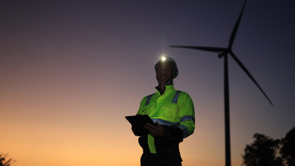 Site engineer with green reflective safety jacket and headlamp working on a digital tablet at a windmill farm © naraichal