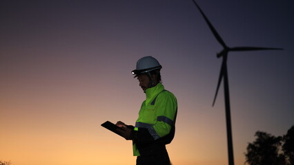 Site engineer with green reflective safety jacket and headlamp working on a digital tablet at a windmill farm © naraichal