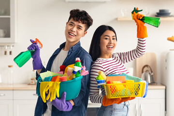 Smiling asian couple posing while cleaning kitchen, holding spray bottles and buskets with cleaning...