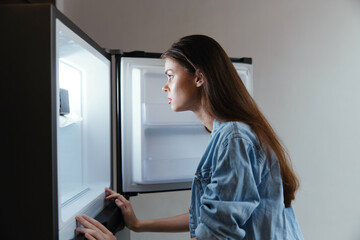 Young female looking into an empty fridge, showcasing emotions of disappointment and contemplation Casual denim jacket, modern kitchen setting, minimalistic design