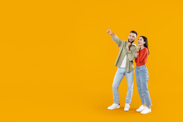 A Caucasian couple stands on a vibrant yellow background, joyfully pointing at copy space and laughing together. Their expressions reflect love and affection