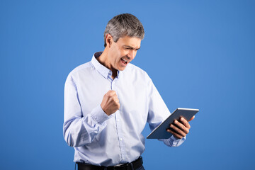 A middle aged man expresses joy and excitement while holding a tablet. His raised fist and...