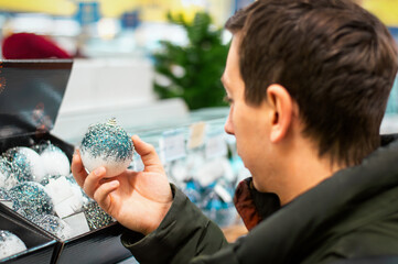 A man chooses white Christmas balls in a store against a blurred background. Christmas shopping concept