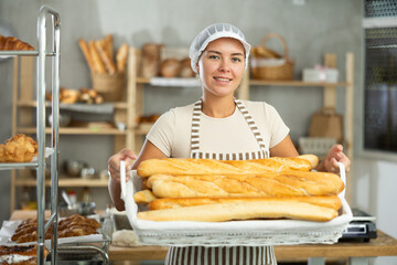 Skilled young female baker in mid-twenties presenting wicker tray of fresh french baguettes with satisfied smile, standing in warm inviting bakery filled with variety of baked goods