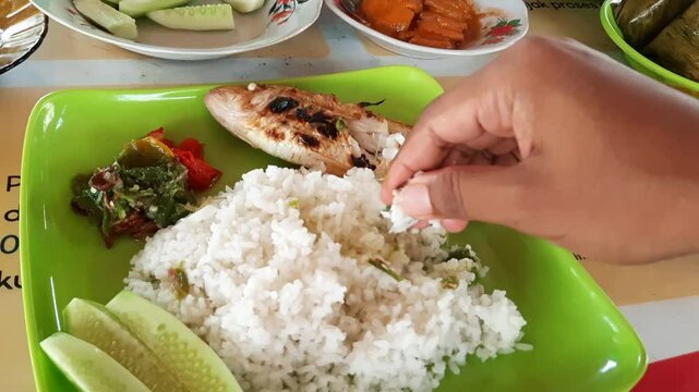 POV of a man hand enjoying Lauak Sambam, a traditional grilled fish dish from Padang Pariaman, Indonesia. Traditional seafood