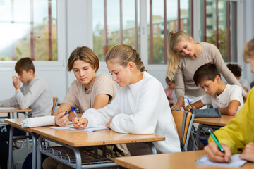 Teenage boy and girl listening to teacher and writing exercises in notebook at lesson in secondary school