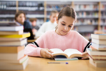 Young girl sitting at table in reading room in library and doing homework.