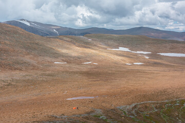 Man near vivid orange tent in sunlit alpine valley against high mountains silhouettes under cloudy sky. Layered mountain dramatic aerial view with sunlight and shadows of clouds on stony rocky pass.