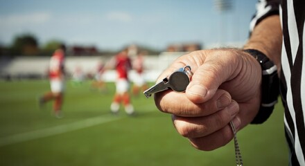 A Referee's Hand Holding a Whistle, with a Blurred Field and Players in the Background During a Football Game
