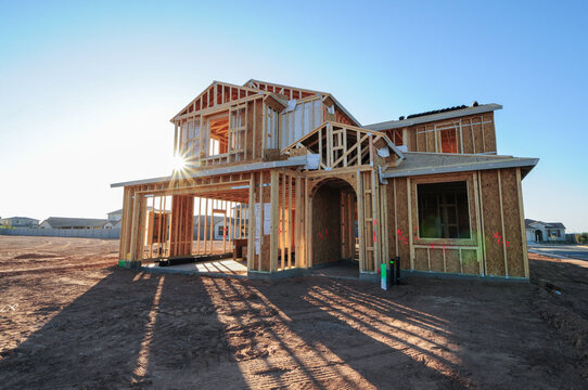 The wooden framing of a single-family, two-story home under construction, with sun rays filtering through the structure, casting diagonal shadows on the brown dirt of the house lot