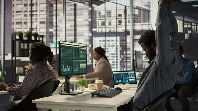Black male CEO dealing with back pain and stretching his muscles at the desk, sitting with an incorrect posture and trying to fix it. General manager working with data analytics. Camera B.
