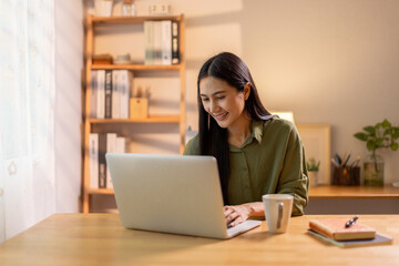 Beautiful young Asian woman working with laptop computer in home kitchen. Work at home. Remote studying E-learning, Watching online education, Webinar at house
