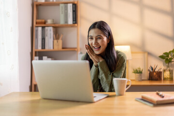 Beautiful young Asian woman working with laptop computer in home kitchen. Work at home. Remote studying E-learning, Watching online education, Webinar at house
