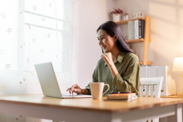 Beautiful young Asian woman working with laptop computer in home kitchen. Work at home. Remote studying E-learning, Watching online education, Webinar at house
