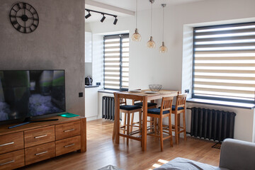 Modern open kitchen interior with wooden high bar table and chairs, sofa and TV, blinds on windows. Stylish living room with dining space. Selective focus.