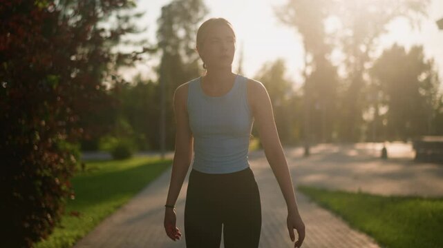 Young lady looking thoughtful while roller skating outdoors on sunny day, swinging her hands for balance, sunlight creating a glowing effect around her, blurred background of lush greenery, trees