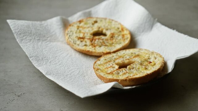 Two toasted bagel halves on paper towel over concrete surface with light shadowing, displaying texture details and crispy surface in a simple culinary setting