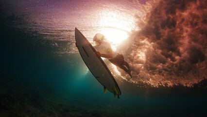 Teen boy surfer dives under the wave with surfing board during sunset