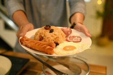 Close up of man holding plate of fried rice with raisins sausage, fried egg and ham slices