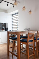 Dining space with wooden high bar chairs, table with white empty plates, glass pendant lights. Modern open kitchen interior. Vertical image.