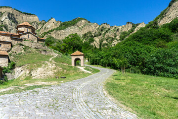 A paved road leads past the refectory to the church of the Shio-mgvime monastery. A rock with cave cells is in the background.