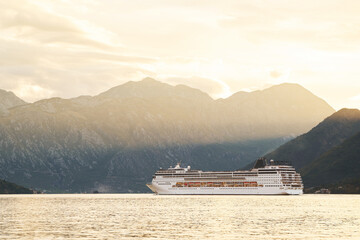 Large white cruise ship sailing on tranquil waters, bathed in the golden light of sunset, against a backdrop of majestic mountains