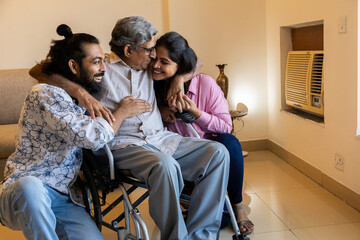 Happy Indian young couple spending time with disabled father on wheel chair at home. Happy parents Day.